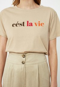 Beige katoenen t-shirt met korte mouwen, bedrukt met "c'est la vie" in bruin, rood en oranje. Gecombineerd met hoge beige broeken.