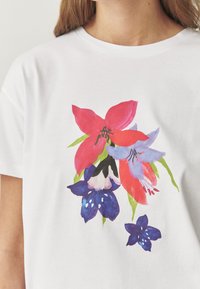 Hvid bomulds T-shirt med farverigt blomsterprint: lyserøde og lilla blomster med grønne accenter, blød tekstur og rund halsudskæring.