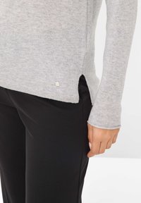 Helles graues Sweatshirt mit langen Ärmeln, abgerundetem Saum, seitlichen Schlitzen und einem kleinen silbernen Akzent am unteren Rand, kombiniert mit schwarzen Hosen.