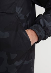 Jachetă neagră cu camuflaj, realizată dintr-un material neted și ușor, cu manșete elastice la încheieturi și o buzunare laterală cu deschidere ascunsă.