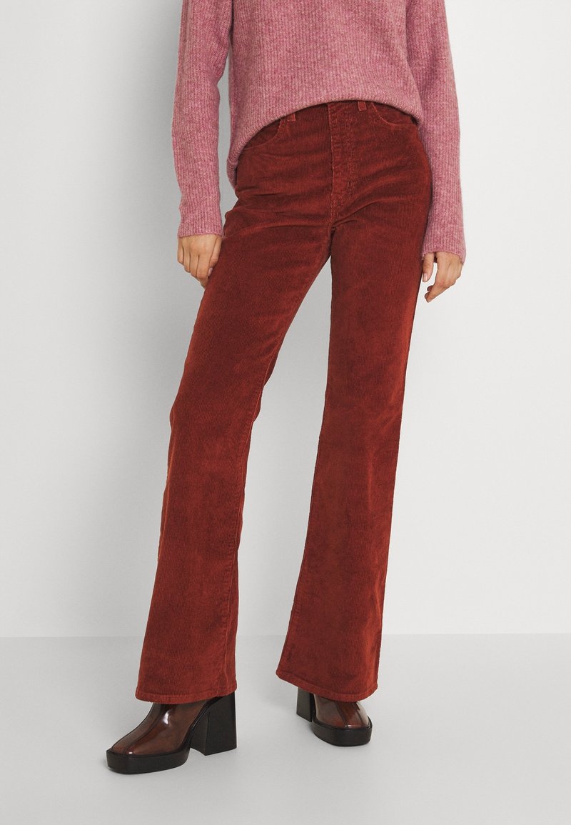 Pantalon évasé en velours côtelé couleur orange brûlé, avec une finition texturée et une taille haute. Associé à un pull en maille côtelée rose et des chaussures à plateforme noires.