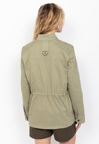 Giacca khaki con vita regolabile tramite cordino, maniche lunghe e una patch con logo sulla parte superiore della schiena, realizzata in un tessuto leggero e morbido.