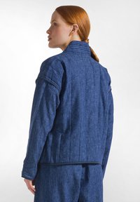 Žena s rudými vlasy svázanými dozadu má na sobě modrou prošívanou bundu a ladící kalhoty, tváří se obrácená doleva na jednobarevném bílém pozadí.