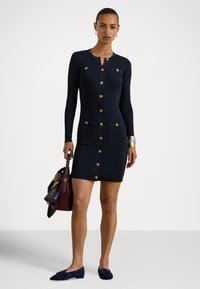 Tumši zila adīta kleita ar riboto audumu, garām piedurknēm, zelta pogām, divām krūšu kabatām un piegulētu siluetu. Kopā ar tumšu somu un loaferiem.