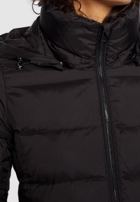 Doudoune noire avec un col haut, dotée d'une fermeture éclair sur le devant et d'accents à cordon de serrage. Tissu lisse et brillant avec des coutures horizontales.