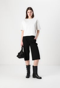 Tricou alb scurt, pantaloni negri largi, cizme negre până la genunchi și o geantă de mână neagră cu model. Texturi netede și design simplu.