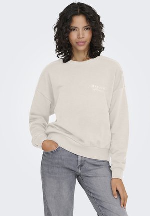 Joven con cabello rizado negro que lleva una sudadera beige con el texto "Blossom" y jeans grises, posando con una mano en el bolsillo.