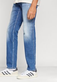 Jeans de denim azul claro con un diseño de corte recto, que presentan un ligero desgastado y detalles de desgaste; combinados con zapatillas deportivas blancas y grises.