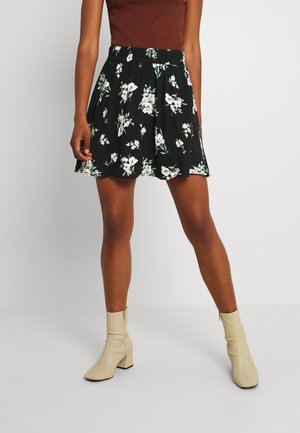 Woman wearing a black floral skirt, brown sleeveless top, and beige ankle boots, standing against a plain light background.