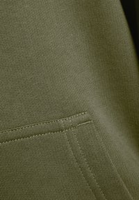 Tessuto verde oliva con una fine trama a maglia, dotato di una tasca obliqua con dettagli di cucitura visibili lungo il bordo.