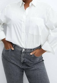 Camisa blanca de botón con mangas largas y un bolsillo en el pecho, combinada con jeans de tiro alto en gris oscuro. Textura de tela suave y corte relajado.