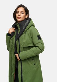Groene parka jack met een zwarte voering en capuchon. Heeft drukknopen en een logo op de kraag. Materiaal is glad met een matte afwerking.