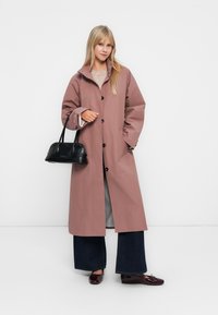Femme portant un long manteau mauve, un pull beige, un jean large foncé, tenant un sac à main noir, se tenant devant un fond blanc uni.