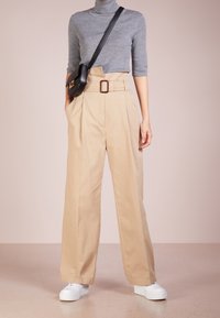 Pantalones de talle alto beige con pliegues, diseño de pierna ancha y un cinturón marrón. Combinados con un suéter de cuello alto gris y zapatillas de plataforma blancas.