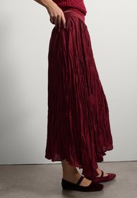 Karmesinrot gefälteter Rock mit strukturiertem Material, breitem Bund und fließender Silhouette. Kombiniert mit burgunderfarbenen, tief ausgeschnittenen Ballerinas.