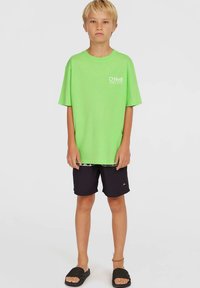 Blonďatý chlapec oblečený v volném jasně zeleném tričku O'Neill, černých šortkách a černých pantoflích stojící na jednobarevném světlém pozadí.