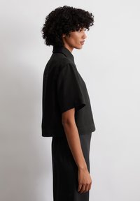 Zwarte korte mouwjacket met kraag, getailleerde pasvorm, gladde stof en strakke lijnen, zijaanzicht tegen een neutrale achtergrond.