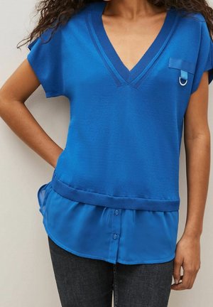 Femme portant un haut bleu à couches avec un col en V, des manches courtes et un onglet de poche décoratif, associé à un jean foncé.