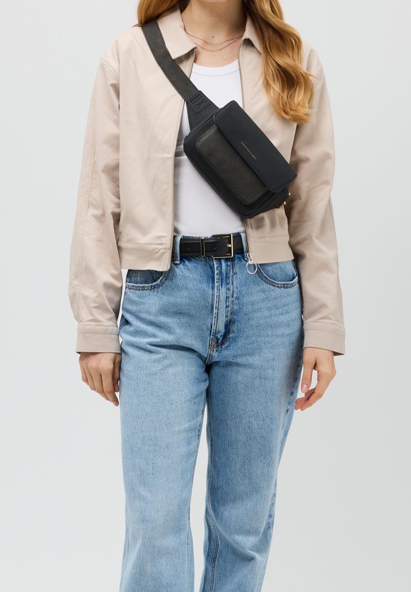 Černá kožená crossbody taška s obdélníkovým tvarem, s klopovým uzávěrem a nastavitelným popruhem, nošená přes béžovou zkrácenou bundu.