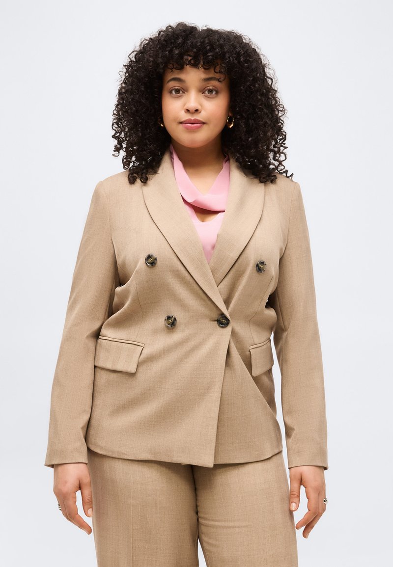 Γυναίκα με σγουρά μαλλιά που φοράει ένα μπεζ διπλόστροφο κοστούμι και ροζ μπλούζα, στέκεται μπροστά σε ένα απλό ανοιχτόχρωμο φόντο.