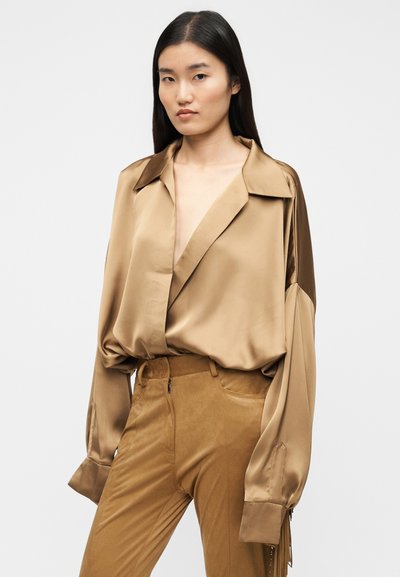 Σατέν μπλούζα σε ανοιχτό καφέ χρώμα με βαθύ V-λαιμόκοψη, φαρδιά μανίκια και αντίθετο σκούρο γιακά, συνδυασμένη με ταιριαστό παντελόνι από σουέντ με στενή γραμμή.