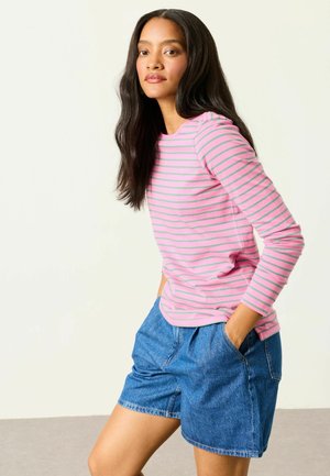 Frau mit langen dunklen Haaren, die ein rosa-grünes gestreiftes Langarmshirt und blaue Jeansshorts trägt, steht mit den Händen in den Taschen.