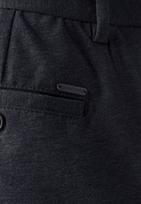 Pantaloni grigio scuro in tessuto strutturato con tasca frontale. Presentano una piccola etichetta con il marchio e una chiusura con bottone.