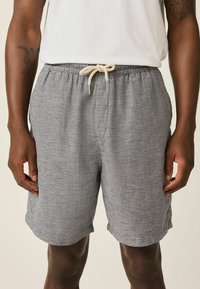 Mann trägt graue, strukturierte Shorts mit Kordelzug und ein weißes Hemd, dargestellt von der Mitte des Oberschenkels bis zur Taille, die Hände entspannt seitlich.