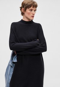 Černé pletené šaty s rolákem, dlouhými rukávy, volným střihem a bočními rozparky; vrstvené s světle modrou džínovou košilí.
