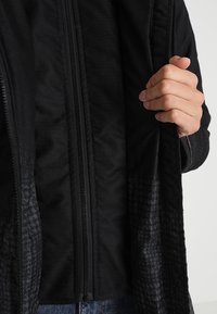 Veste noire avec des accents en tissu texturé, dotée d'une fermeture éclair à l'avant et d'une doublure intérieure. Le matériau présente une finition lisse et un motif subtil.
