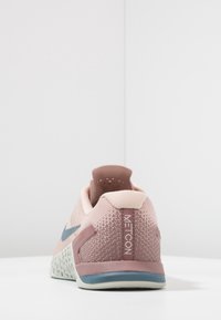 Zapatilla deportiva de color rosa claro con parte superior de malla texturizada, lengüeta en el talón y suela gris con un agarre con patrones. Marca "METCON" en el talón.