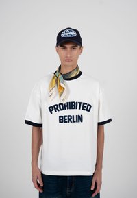 Jeune homme portant un t-shirt blanc "Prohibited Berlin", une casquette bleu marine avec le même texte, et une écharpe colorée à motifs nouée autour du cou.