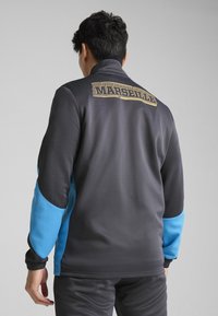 Grå och blå träningsjacka med dragkedja i kragen. "MARSEILLE" i guld över ryggen. Slät textur, figursydd design, elastiska ärmslut.