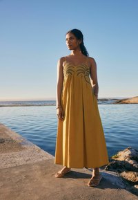 Robe maxi jaune avec une broderie raffinée au niveau du corsage, des bretelles fines et une coupe ample. Portée avec des sandales plates tan près de l'eau.