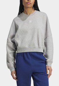 Grå kortärmad sweatshirt med rundad hals och långa ärmar, prydd med en liten vit logotyp på bröstet, i kombination med blå byxor.