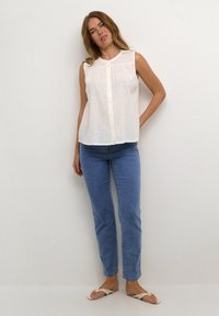 Blouse blanche sans manches avec un devant à boutons, fabriquée dans un tissu texturé. Associée à un jean bleu à jambes droites et des sandales de couleur claire.