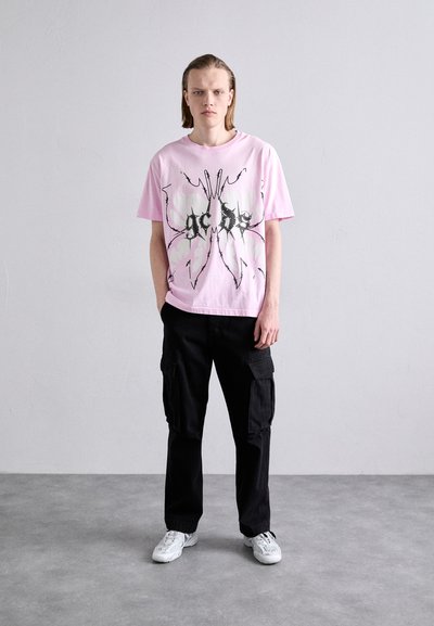 T-shirt rose à manches courtes avec un design graphique noir, associé à un pantalon cargo noir et des baskets blanches, sur un fond neutre.