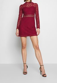 Burgunderfärgad spetsklänning med långa ärmar och en åtsittande passform. Har en genomskinlig ok och utskärningar vid midjan. Kombinerad med svarta remsandaler.