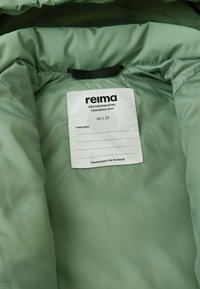Interiorul jachetei verzi impermeabile cu un etichetă textilă care afișează "reima," mărimea 92/2T, și o secțiune de instrucțiuni de întreținere scrise de mână.