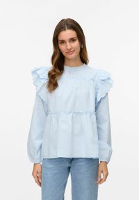 Blusa azzurra chiara con maniche a ruffle, strati arricciati e un collo alto. Il tessuto sembra liscio, con una leggera lucentezza e polsini elasticizzati.