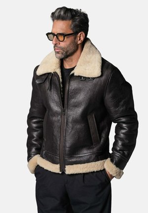 Homme portant une veste en cuir marron foncé avec col et poignets en shearling crème, pantalon noir et lunettes, regardant sur le côté les mains dans les poches.