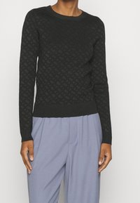 Pull noir en maille avec un motif en relief en forme de losange, col rond et poignets côtelés. Porté avec un pantalon gris clair.
