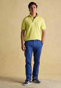 Žlté polo tričko s krátkym rukávom a golierom, skombinované s modrými nohavicami. Model má na sebe námornícke tenisky s bielymi pruhmi. Neutrálny pozadie.