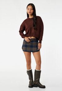 Brązowy dzianinowy sweter cropped, niebieska kratkowana mini spódniczka z paskiem oraz brązowe kozaki do kolan z kremowymi mankietami i akcentami klamrowymi.