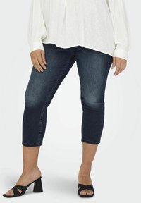 Dark wash denim capri jeans with a slim fit, cuffed hem, and mid-rise waist, paired with open-toe black block-heel sandals.