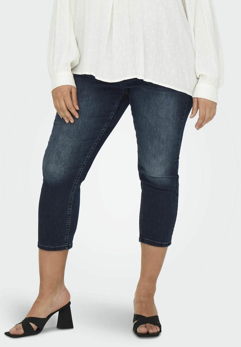 Dark wash denim capri jeans with a slim fit, cuffed hem, and mid-rise waist, paired with open-toe black block-heel sandals.