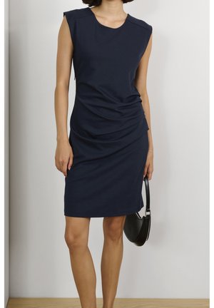 Femme portant une robe bleu marine sans manches jusqu'aux genoux, tenant un petit sac à main noir, debout contre un mur blanc et un sol en bois.