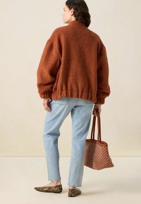 Veste surdimensionnée en rust avec un col haut, associée à un jean droit bleu clair. Le modèle porte un sac marron texturé.