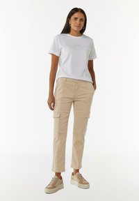 Witte katoenen t-shirt met "VOILA" gedrukt in goud, gecombineerd met tan kleurige cargobroek met zijzakken, en beige platform sneakers.