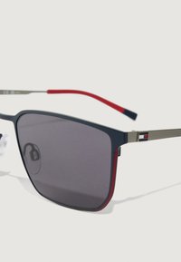 Ματ μαύρα και κόκκινα ορθογώνια γυαλιά ηλίου με σκούρους φακούς και λεπτούς χρυσούς μεταλλικούς βραχίονες που διαθέτουν μια μικρή λεπτομέρεια σημαίας.